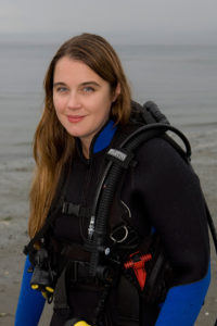 Instructor and cofounder Amanda Castleman, a Caucasian lady with long brown hair, seen in cold-water dive gear against a misty Pacific Northwest oceanscape. Photo by Mike Keran.