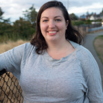 Instructor and founder Naomi Tomky, a Caucasian woman in a grey shirt, smiling as she leans against a fence in the Pacific Northwest. A curving trail and evergreen trees are visible in the background