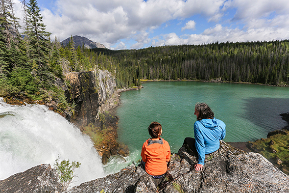 Two women sitting at the edge of a Canadian Rockies cliff in Tumbler Ridge, BC, right beside a waterfall dropping into a blue-green alpine lake