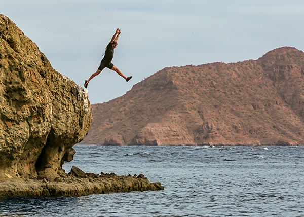 A dude in a tropical shortie wetsuit jumping off a sea cliff in Baja, Mexico, with ruddy mountains in the background