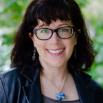 Instructor Alex Leviton (a white woman with brown bangs and shoulder-length hair, glasses, a bright smile and pretty blue necklace. You can see PNW foliage in the background.)