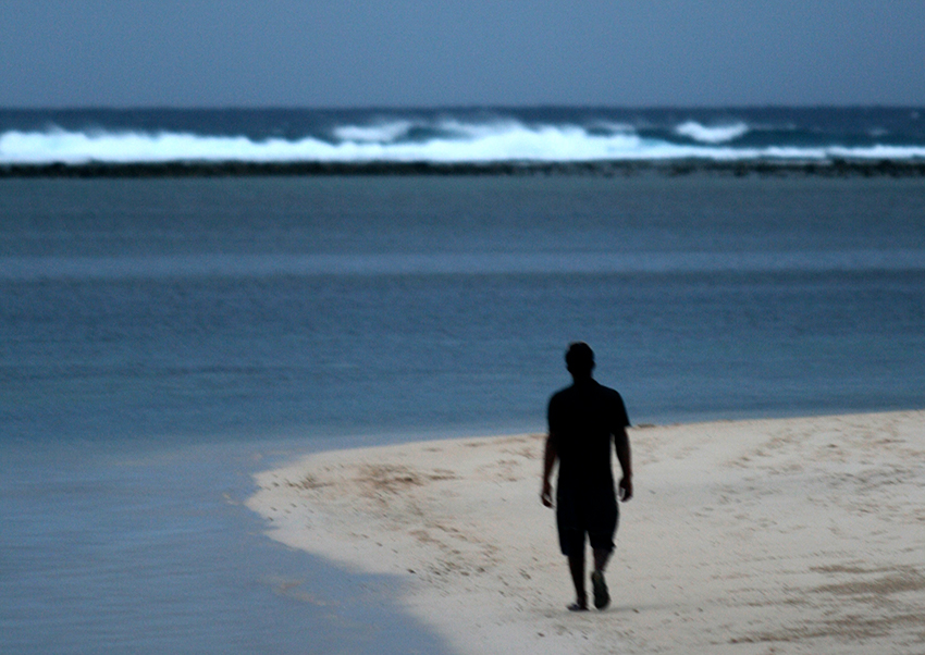 The silhouette of a man walking down a tropical white beach in the Cook Islands, facing a story sea and reef break