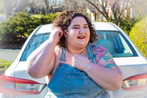 Chaya Milchtein, an automotive writer and plus size model sits on the trunk of a white car on a sunny day. She is tucking her dark curls behind one ear and a colorful tattoo sleeve peeks out from under her overalls. She looks playful and welcoming.