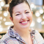 Instructor Naomi Tomky, a smiling brunette Caucasian lady with a casual up-do and a pale white-and-purple plaid shirt. The background shows fairy lights, blurred by bokeh