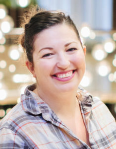 Instructor Naomi Tomky, a smiling brunette Caucasian lady with a casual up-do and a pale white-and-purple plaid shirt. The background shows fairy lights, blurred by bokeh