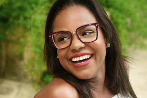 A brown-skinned woman glances off-camera with a happy smile. She has dark hair parted in the middle and dark-framed glasses on. Greenery is visible in the background.