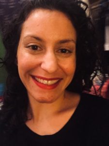 A close-up of a Latinx woman with dark hair, a wide smile and red lipstick. She's wearing a black v-necked shirt.