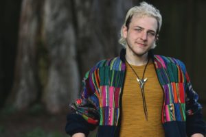 Kurt Suchman (they/them), a white person with bleach blonde hair, blue eyes and a smile with some 5 o'clock shadow. Kurt is wearing a patchwork jacket and mustard-colored T-shirt with a bulls-head bolo tie. What appears to be a large tree appears in shawdowy bokey behind them.