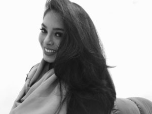 A black-and-white shot of writer Shaistha Khan, a second-generation Indian living in Saudi Arabia. She is peering out from under a swoop of long, lustrous dark hair with bright eyes and is smiling. She appears to have a pashmina on and is posed against a white background.