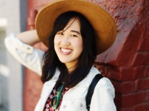 Writer Julia Chen, a Chinese-American woman smiling, against a dark red brick background. She's gripping a straw hat and is wearing a colorful top under a summery white shirt. Her black hair is shoulder-length