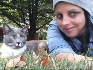 Sara Giza, a writer and activist. Pic: a smiling Polish-Syrian woman sprawled in the grass. She's wearing a powder blue hat and shirt, and is featured with a gray and white cat. 