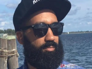 Carlos Matias, a second-generation Dominican-American food writer. He's wearing a dark baseball cap and sunglasses, and has a beard and mustaches. He appears to be standing on a dock with dark-blue water behind him.