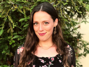 Bogotá-based journalist Jennifer Bitterly, a young white woman with long brown hair, a black dress with a subdied flower print and a kind smile. She's standing in front of an evergreen dappled by the sun.