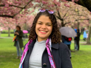 A smiling Latinx woman with curly, dark shoulder length hair, and a fuscia scarf and sunglasses that compliment the cherry blossoms behind her