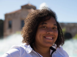 Lauren J. Mapp, a Black-Indigenous journalist, smiling, with curls reaching to the collar of her white button-down shirt. She's outside, against a sunny background of trees, a bridhe and maybe a golden-hued stone church?
