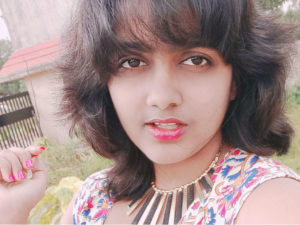 Sakshi Udavant, an Indian journalist, with fashionable bangs and shoulder-length dark hair. She's wearing a brightly patterned top that matches her nails and lipstick. She's outside in a verdant yard with a stucco building in the background.