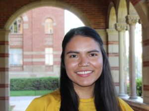 Mexican-American writer Rocio Sanchez-Nolasco, a smiling young Latinx woman with long dark hair, pulled back on top, and a canary-yellow shirt. She's standing in a portico with hedges, red-brick arches and marble columns in the background