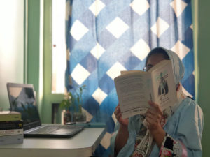 A writer in a hijab reading a book. Tasneem sits at a desk with a laptop in front of a sunlit blue-checkered drape