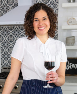 Food writing instructor Kae Lani Palmisano, a smiling biracial woman with dark shoulder-length curls. She's posed in a chic kitchen, holding a glass of red wine and wearing a white, short-sleeved blouse with a pussycat bow.