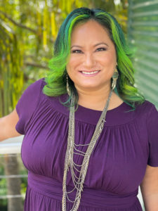 A smiling Asian-American woman with lime-green shoulder-length hair and silver necklaces draping over a purple short-sleeved top. The background looks rainforesty.