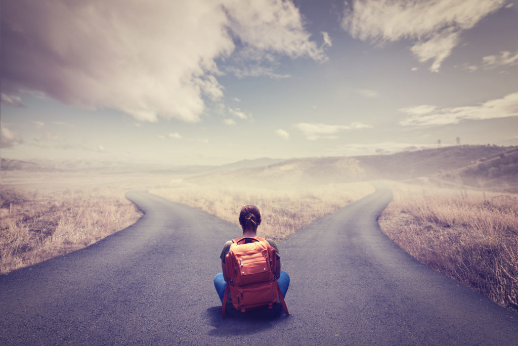 A woman with a red backpack is seen from behind, sitting cross-legged where a road divides. Dry grass stretches on both sides and mountains can be seen in the distance. The faded-blue sky has fluffy clouds in it. (Image by Sondem via Shutterstock)