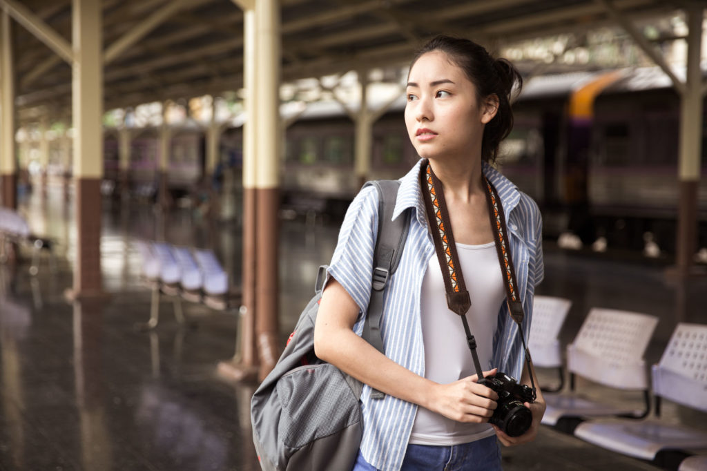 A young Asian woman pauses in a train station. She's carrying a rucksack and camera.