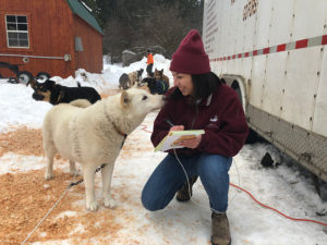 A Mexican-American journalist kneels in the snow while reporting on the Iditarod. She's wearing a purple hat, heans and a purple coat, and holding a notebook. An adorable white husky peers into her face.