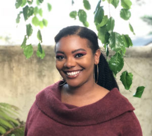 Portrait of a smiling Afro-Creole woman, wearing a burgundy cowl-neck top. Her long straight hair is pulled back on top. Leaves drape near her face and there might be fields in the blurred-out background.