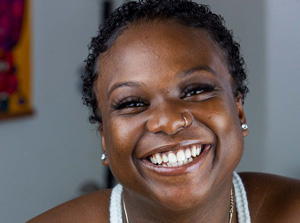 A portrait of a smiling Black woman with short-cropped curls and what appears to be a crocheted white halter top