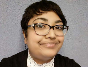 A portrait of an Indo-Trinidadian writer and social worker. She has straight, short-cropped hair and black-framed stylish glasses on.