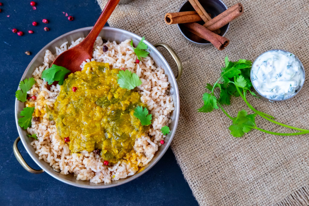 A rice dish with a green-yellow curry in a silver-looking round bowl. It stands on a burlap placemat, which also holds some herbs and cinnamon sticks