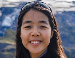 A smiling Malaysian writer with long, dark hair worn loose and sunglasses on her head. She stands in front of a mountain scene with what appear to be glaciers