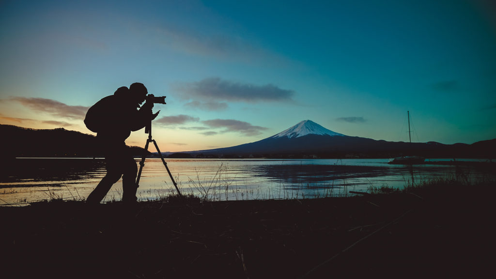 Silhouette of a landscape photographer with a backpack and a camera on a tripod. It’s twilight. A lake and snow-gilded stratovolcano dominate the background.