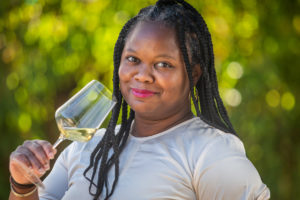 A smiling Black woman with long black braids, a while long-sleeved tee and she's holding a wine glass near her mouth and is wearing fuchsia lipstick. Sunlit greenery is boca-ed behind her