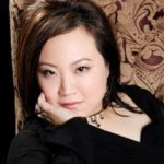 The author Leslie Hsu Oh, a woman of Chinese descent. She has side-swept dark brown hair, a black top and choker with black beads dangling. Leslie's leaning on one hand with a beige and brown brocade behind her. She looks serene and thoughtful.