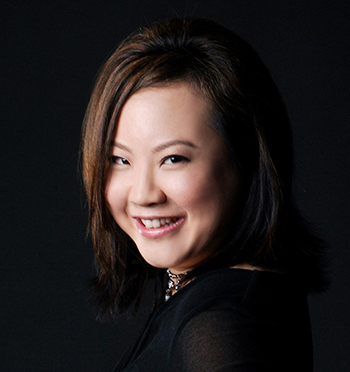 The author Leslie Hsu Oh, a woman of Chinese descent. She has side-swept dark brown hair, a black top and choker with black beads dangling, and the background is also black. She's smiling.