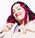 Image of Instructor Naomi Tomky, a white Millennial against a white background. She has fuschia streaks in her dark shoulder-length hair, a wry smile, and is holding a light pink fluffy fleece with two hands near its labels.