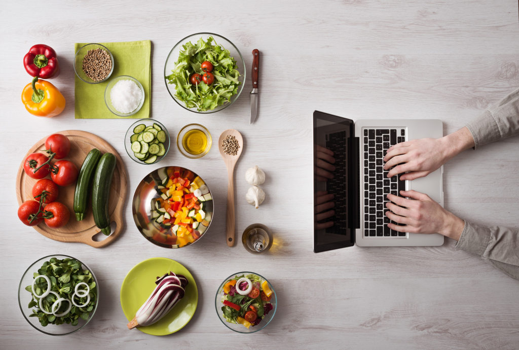 Licensed Shutterstock image by Stock-Asso. It's an overhead shot of a white-grey marble countertop. White hands rest on a Mac-looking laptop on the right side of the frame. The rest is mise en place bowls with radicchio, green lettuce shredded, bell peppers, etc.