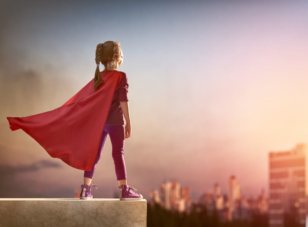 A dreamy shot of a mid-sized girl —wearing a red cape — overlooking a distant, sunset-soaked city.
