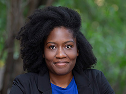 Portrait of a Black musician and writer. She's portrayed outside against a bokehed forest background. Her natural hair is swept to one side and reaches her shoulders. She's smiling slightly and wearing a blue top and black blazer.