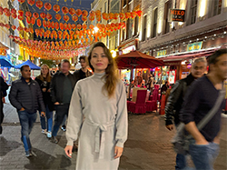 Portrait of a writer standing in what appears to be a night market, with strings of Chinese lanterns above. The crowd blurs past her, as she stares directly into the camera. She's wearing an elegant light grey tie-front dress and has her long blonde-brown hair swept over one shoulder.