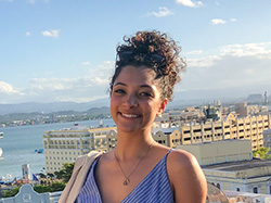 Portrait of a Latinx writer. She's wearing a blue-and-white striped sundress with a V-neck and spaghetti straps, sitting on a scenic overlook with a Mediterranean-looking harbor behind her. Carolina's dark, curly hair is piled on top of her head, along with a pair of sunglasses.