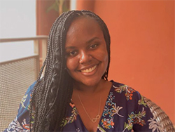 Portrait of a Black Haitian-American writer. She's smiling with her long box braids parted to one side, cascading over the shoulder of her v-necked floral blouse (navy background, white and pink pattern). She's illuminated softly by a window and the wall behind is a warm pink-orange tone.
