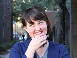 Portrait of a white writer, smiling outside with her hand to her chin and her gaze up and off to the side. She has a mischievous grin and dark brown bangs swept to one side. Her hair appears to be in a bob, and she's wearing a purple sweater over a grey V-neck. She seems to be on an urban sidewalk.