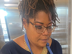 Portrait of a Black writer making empanadas (the food is out of the frame). Her thin box braids are piled on top of her head, and she's looking down through glasses. She's wearing a thick gold necklace, a navy V-neck shirt and a denim-colored apron.
