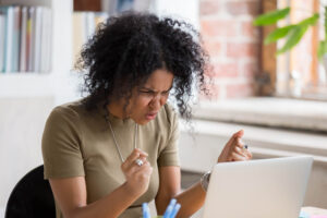 A woman who appears Black shakes her hands at a laptop. She has springy curls to her jawline and is wearing a beige shirt. Behind her you can see a bookshelf, window and brick wall