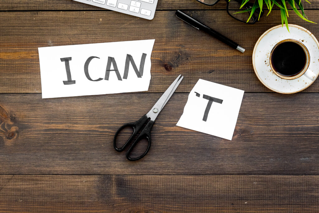 Stock art of a rustic wooden table, shot from above. On it, a pair of scissors separates two piece of paper, one reading "I can" and the other "'t". A succulent and tea cup are visible top-right to set the scene