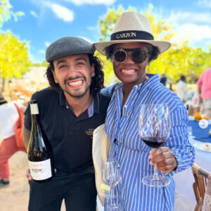 Two friends pose in a vineyard. On the right, J'nai, a Black wine writer, wears a blue-and-white=striped shirtdress and a straw boater with a black ribbon printed with "Garzon." To the left is legendary winemaker Alberto Antonini holding a bottle. He's wearing black jeans, a black polo and a grey cap that looks felty. They appear to be in a festive outdoor setting with trees glowing line-chartreuse in the sunlight. Clouds lightly streak the blue sky.
