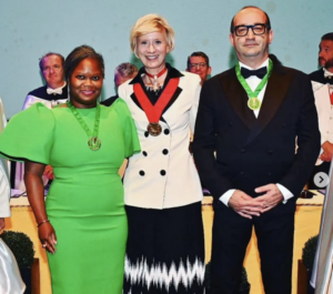 Three people stand before a banquet table. Two are white: the man wears a black suit and the blonde woman a white top and black skirt. To the left is J'nai, a beautiful Black woman. She's wearing a lime-green dress with massive shoulders and has a "knight of champagne medal around her neck