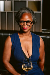 J'nai Gaither, a Black wine writer with curly hair, faces the camera smiling. She's wearing a navy blue sleeveless dress, black glasses and red lipstick, and is holding a glass of champagne.